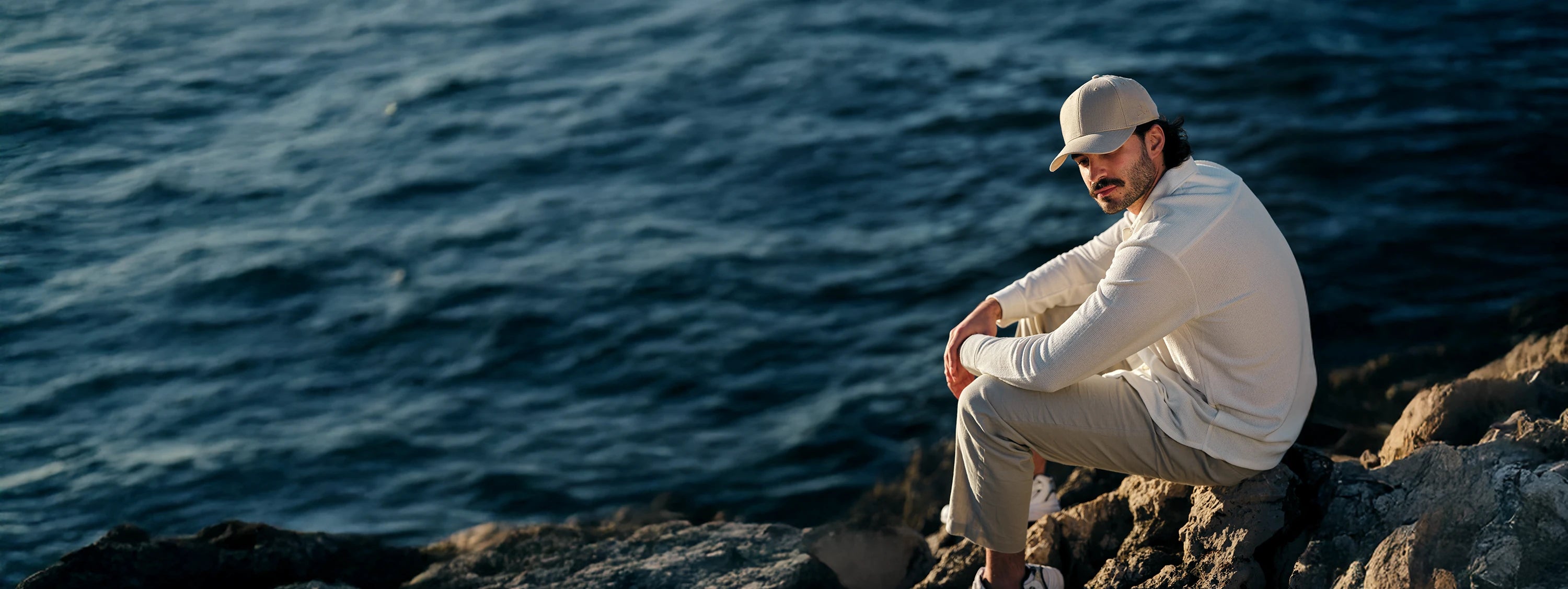 Homme portant une casquette ADN assis sur des rochers au bord de l'eau en tenue décontractée