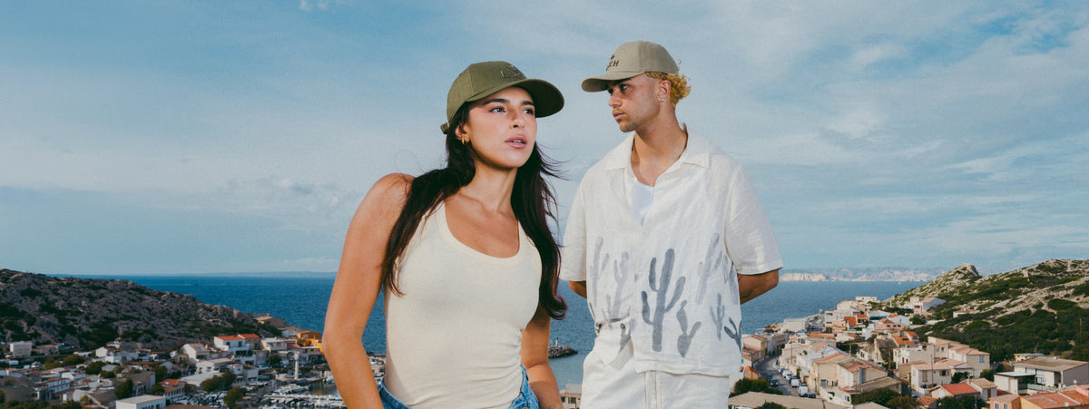 Couple portant des casquettes LXH kaki et beige au bord de la mer, collection printemps été 2026.