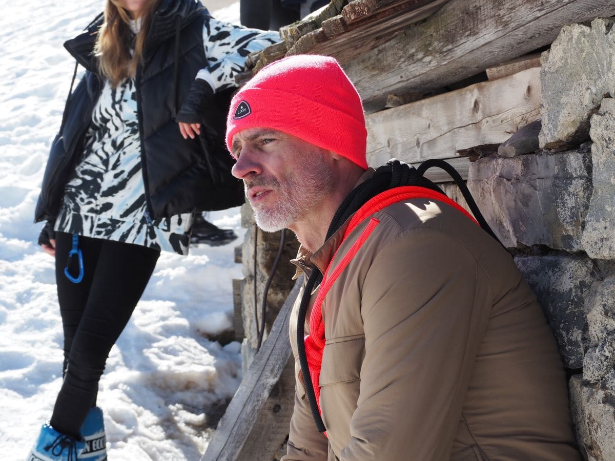 Homme portant un bonnet acrylique rose, assis sur la neige avec un style hivernal