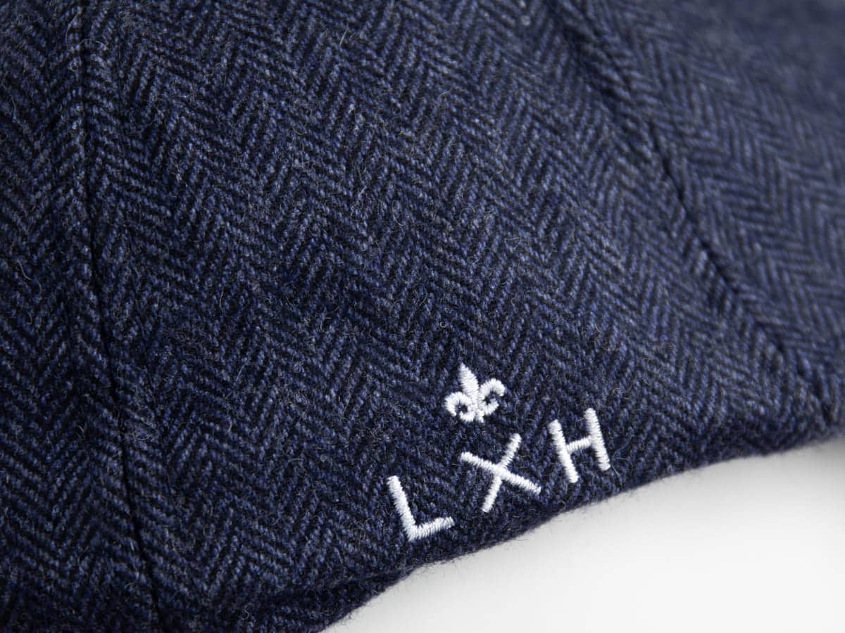 Macro du tissu chevron bleu marine du béret LXH avec broderie blanche du logo fleur de lys et LXH en gros plan.